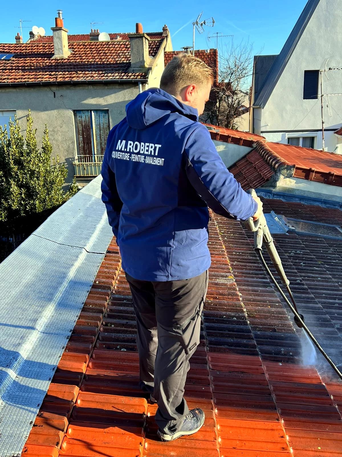 Un couvreur sur une toiture, lance de nettoyage en main, mousse encore visible sur les tuiles